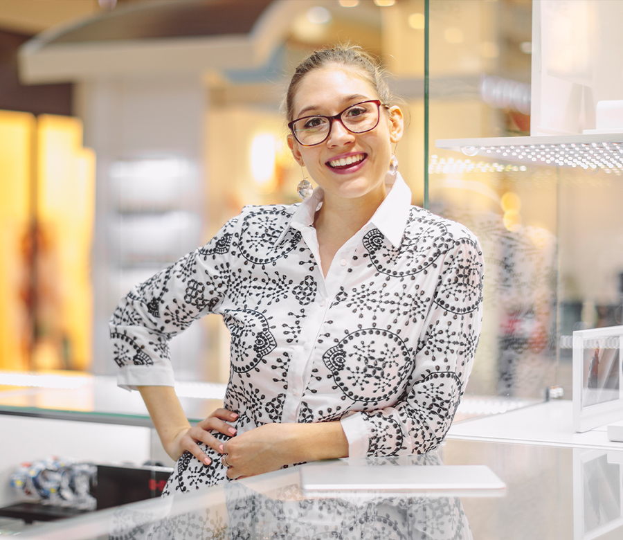 Employée de magasin souriante, portant des lunettes et une chemise blanche à motifs noirs, derrière un comptoir en verre dans un centre commercial.